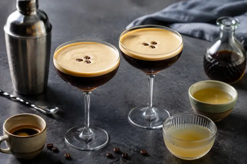 Köpüklü espresso martini ve alkolsüz kahve kokteyli; yanlarında kokteyl shakerı, espresso, kahve şurubu ve aquafaba.